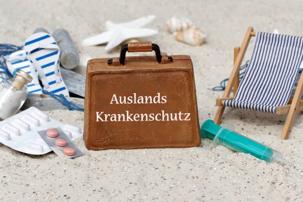 Ein Koffer mit der Schrift "Auslands-Krankenschutz" liegt am Strand mit Medikamenten und Liegestuhl