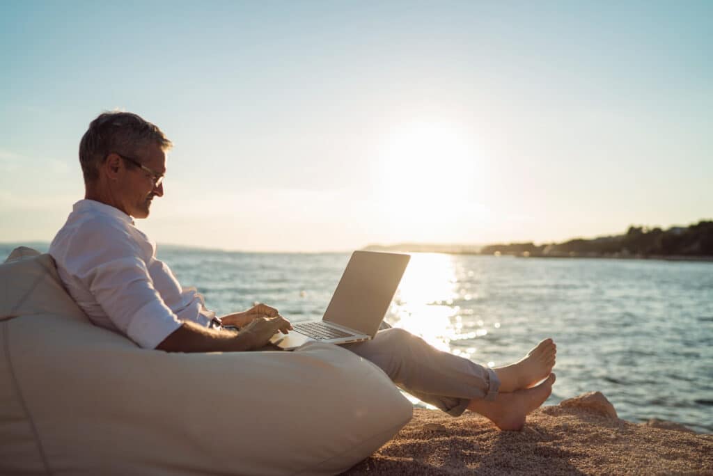 Mann sitzt am Wasser und arbeitet an seinem Laptop