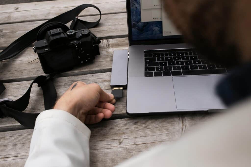 Mann steckt eine SD-Karte in einen Multi-Port-Adapter, der mit einem Laptop verbunden ist, während eine Kamera auf einem Holztisch daneben liegt.